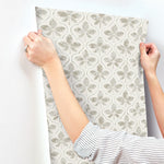 A person with a striped shirt is applying a sheet of Sevilla Onyx Wallpaper Black, Brown (60 Sq.Ft.) by York Wallcoverings to a white wall. The wallpaper, part of the SureStrip collection, features a design of light beige and gray clover-like shapes in a repeating pattern, adding geometric elegance to the room.