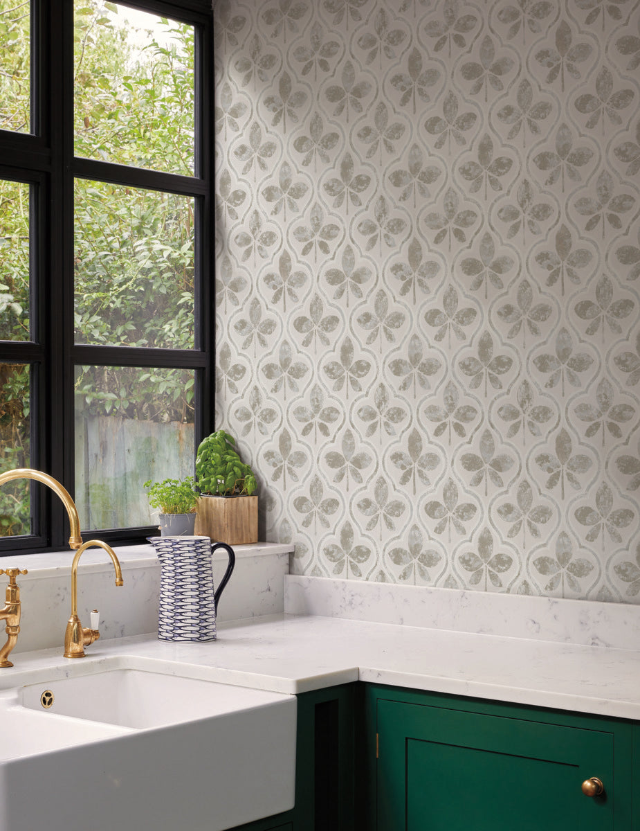 A kitchen corner with a white marble countertop, a white farmhouse sink, and a brass faucet. The walls feature York Wallcoverings' Sevilla Ochre Wallpaper Yellow (60 Sq.Ft.) with green and white geometric elegance. A potted basil plant and a striped jug grace the counter. Large windows let in natural light and offer a garden view.