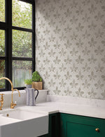 A modern kitchen with white marble countertops, a green cabinet, and a white sink with a brass faucet. The wall features a Sevilla River Rock Wallpaper Grey (60 Sq.Ft.) by York Wallcoverings in light gray and white tiles, and there's a large window with black frames letting in natural light. A potted plant and a striped jug sit on the counter.