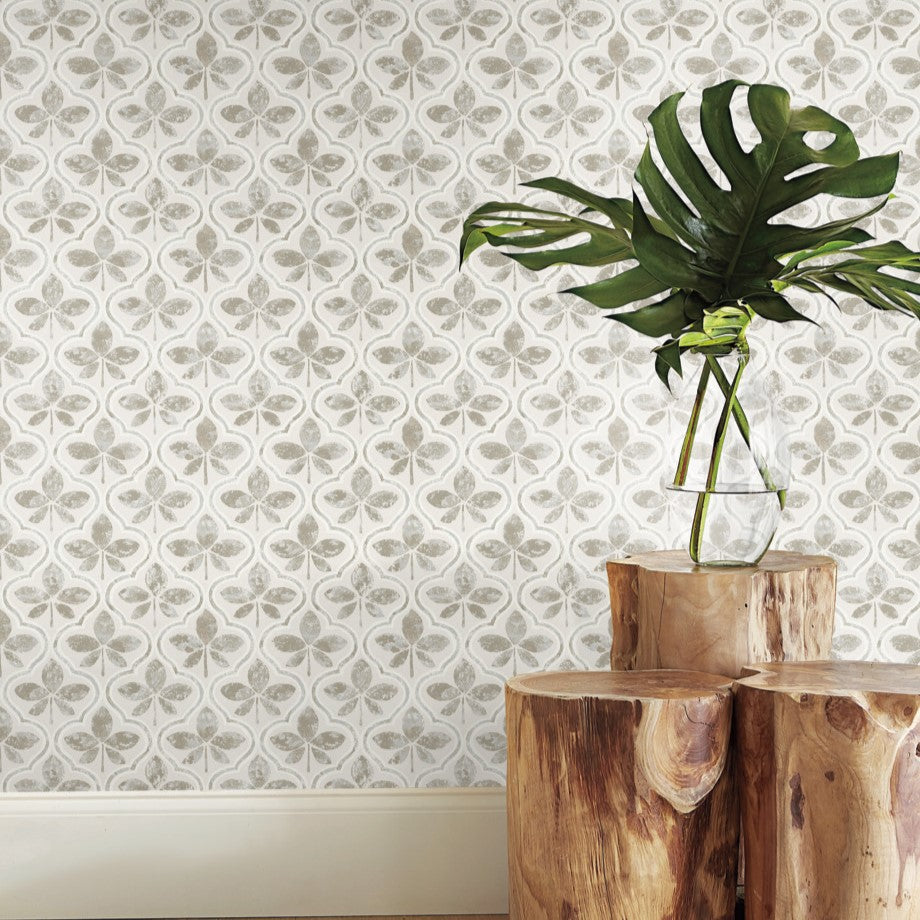 A clear glass vase holding a large green monstera leaf is set on a stack of three wooden logs arranged as a small table. The background features a wall adorned with York Wallcoverings' Sevilla Ochre Wallpaper Yellow (60 Sq.Ft.), showcasing light gray floral and clover patterns that elegantly complement the scene.