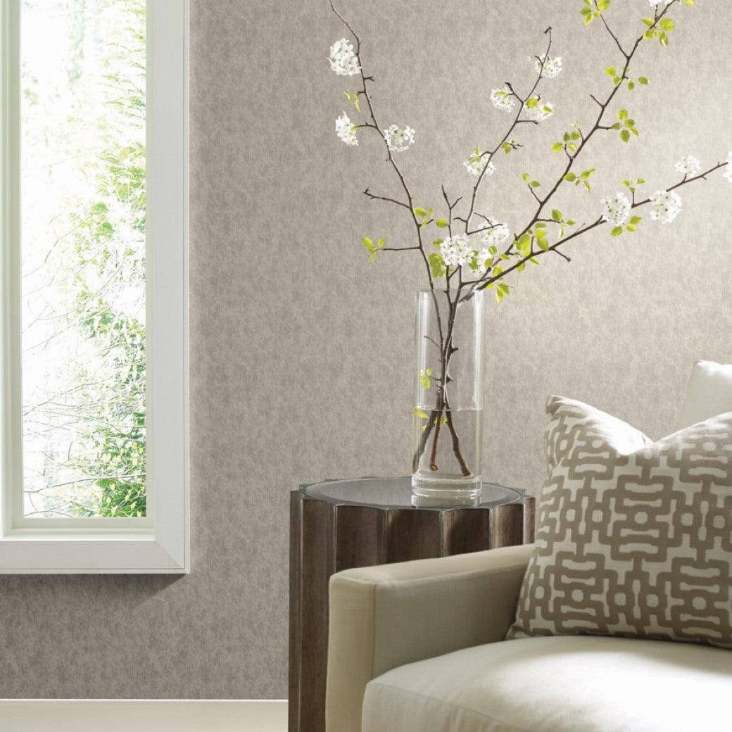 Living Room - A minimalist corner features a light beige sofa with a patterned cushion against York Wallcoverings' Silver on Grey Shimmering Patina Wallpaper. A small wooden side table holds a tall vase with blossoming branches, while soft natural light casts gentle shadows through the window