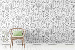 A minimalist room featuring a wooden chair with a green cushion against a white Decor2Go Wallpaper Mural with a black doodle pattern of various whimsical flora and fauna.