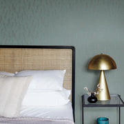 Bedroom - A modern showcases a rattan headboard, white pillows, and a neutral bedspread. A black side table from our Modern Metals Collection holds a gold lamp and decor. Smokey Blue & Silver Sprigs Wallpaper by York Wallcoverings features a leafy pattern in soft green