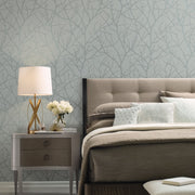 Bedroom - A stylish showcases a beige tufted headboard with neatly arranged pillows on a neutral-toned bed. A wooden nightstand holds a modern lamp and a vase of white flowers, while York Wallcoverings' Smokey Blue & Silver Trees Silhouette Wallpaper adds elegance with its subtle branch pattern
