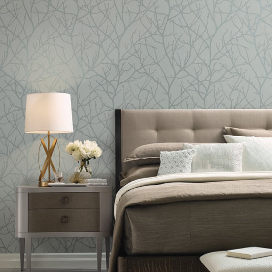 A stylish bedroom showcases a beige tufted headboard with neatly arranged pillows on a neutral-toned bed. A wooden nightstand holds a modern lamp and a vase of white flowers, while York Wallcoverings' Smokey Blue & Silver Trees Silhouette Wallpaper adds elegance with its subtle branch pattern.