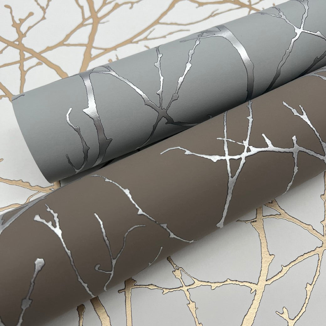 Two rolls of York Wallcoverings' Smokey Blue & Silver Trees Silhouette Wallpaper (57 SqFt) feature a metallic branch design. One roll is gray, the other brown, both with silver accents, on a surface reflecting their pattern with a gold branch silhouette.