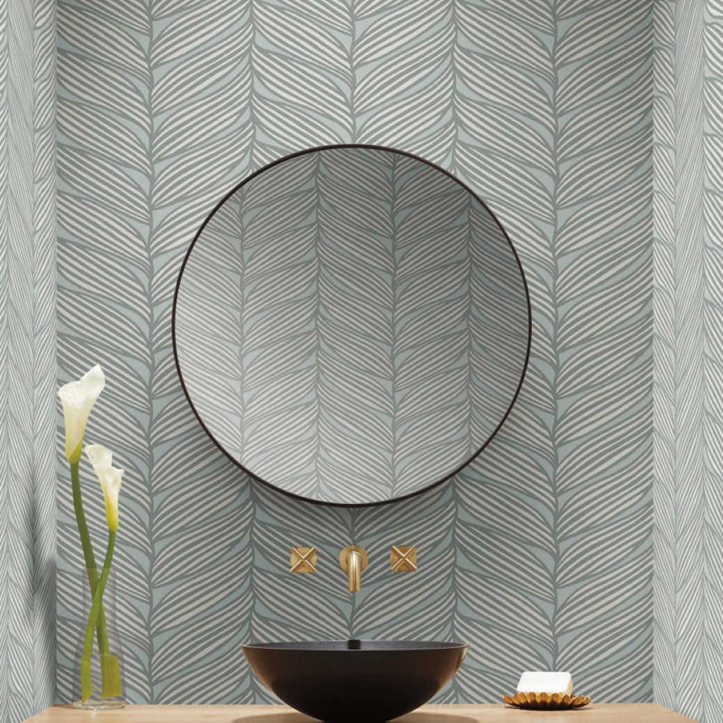 The bathroom features a circular mirror above a black basin with a gold faucet from the Modern Metals series. The walls display Spa & Silver Luminous Leaves Wallpaper by York Wallcoverings. A vase with white flowers and a soap dish adorn the wooden counter for added modern elegance.