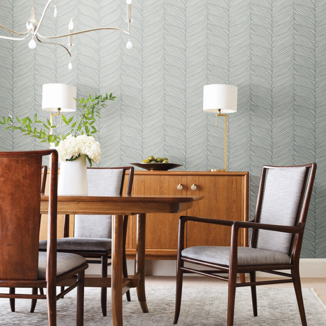 A dining room with a wooden table and chairs, a modern light fixture from the Modern Metals series, and a mid-century sideboard. York Wallcoverings' Spa & Silver Luminous Leaves wallpaper decorates the wall. Two lamps and a vase with palm leaves sit on the sideboard, while a rug covers the floor.