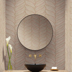 A modern bathroom has a round mirror with a black rim against York Wallcoverings' Blush & Gold Luminous Leaves Wallpaper. Below is a black basin with gold faucets from the modern metals collection, completed by a vase with two white flowers and a soap dish.