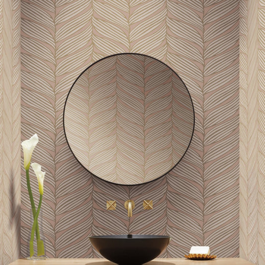 A modern bathroom has a round mirror with a black rim against York Wallcoverings' Blush & Gold Luminous Leaves Wallpaper. Below is a black basin with gold faucets from the modern metals collection, completed by a vase with two white flowers and a soap dish.