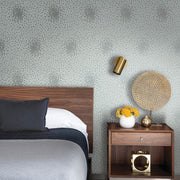 Bedroom - A stylish includes a wooden headboard bed with gray-black bedding, a wooden nightstand with yellow flowers and a lamp. The walls are adorned with York Wallcoverings' Spa & Silver Petite Leaves Wallpaper featuring subtle floral designs, complemented by a gold wall lamp