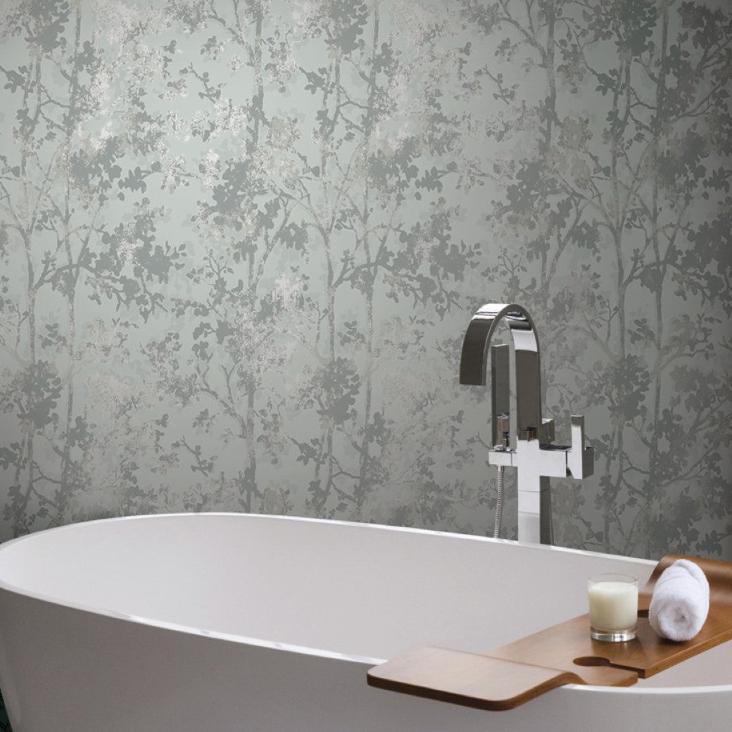 A bathtub with a pewter metallic faucet features a wooden caddy holding a candle and rolled towel, set against York Wallcoverings' Spa & Silver Shimmering Foliage Wallpaper (57 SqFt), showcasing a green tree silhouette pattern.