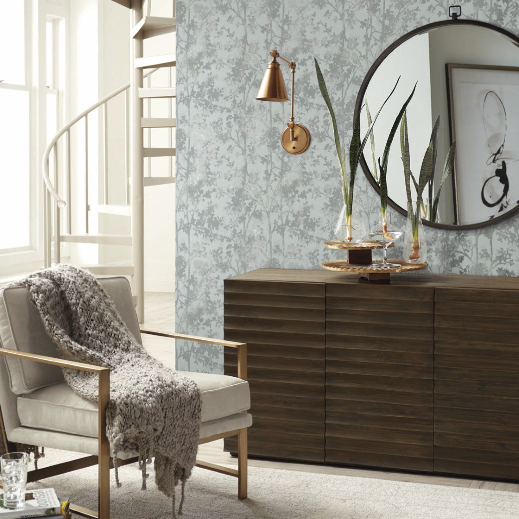 Living Room - A cozy features a silver metallic armchair with a knitted throw. A wooden sideboard rests against York Wallcoverings' Spa & Silver Shimmering Foliage Wallpaper, topped with decor and a round mirror. A copper sconce lights the space, while a spiral staircase is nearby