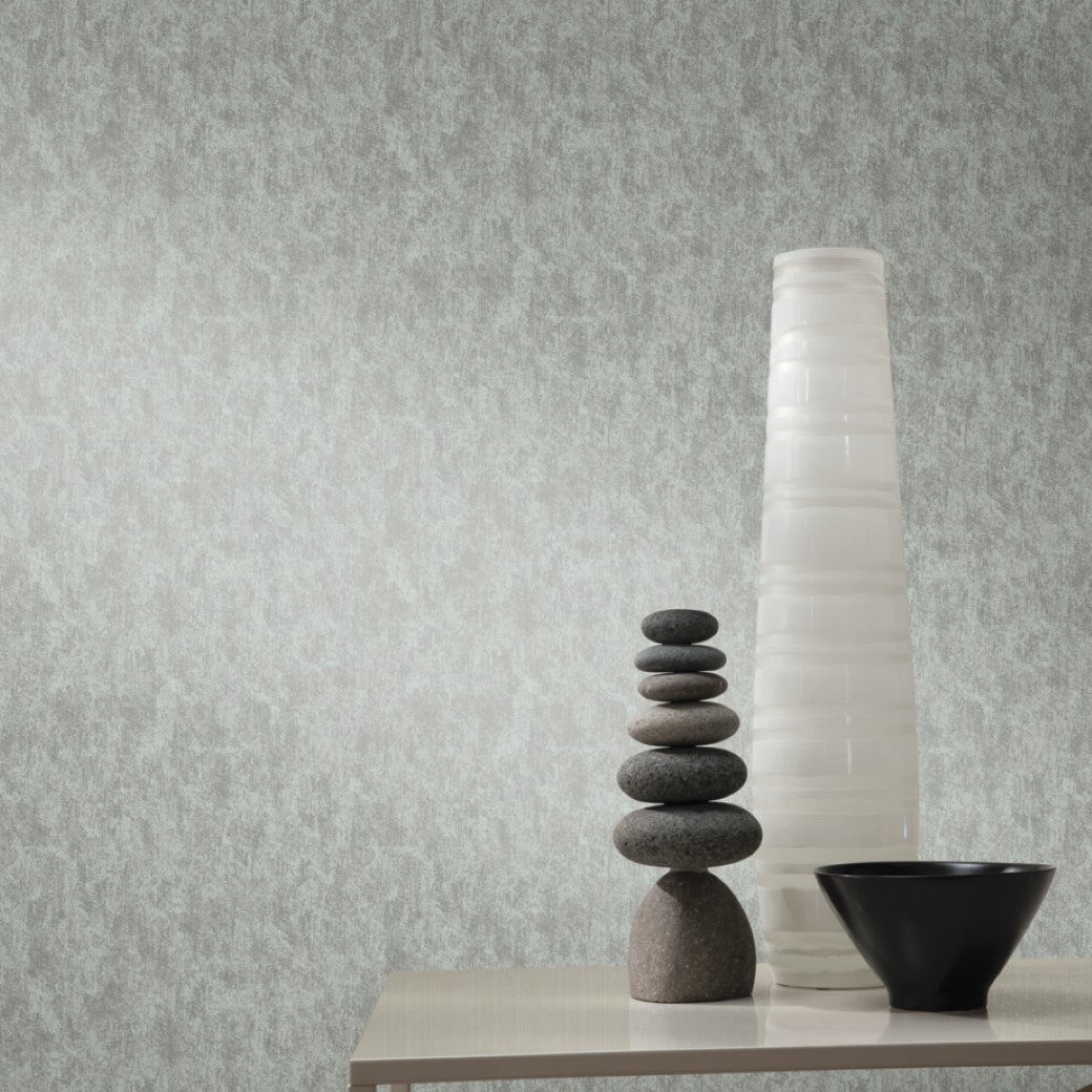 A minimalist table setting includes round stones, a tall vase with horizontal lines, and a small black bowl. The backdrop is York Wallcoverings' Spa & Silver Shimmering Patina Wallpaper, enhancing the textured light grey wall with subtle elegance.