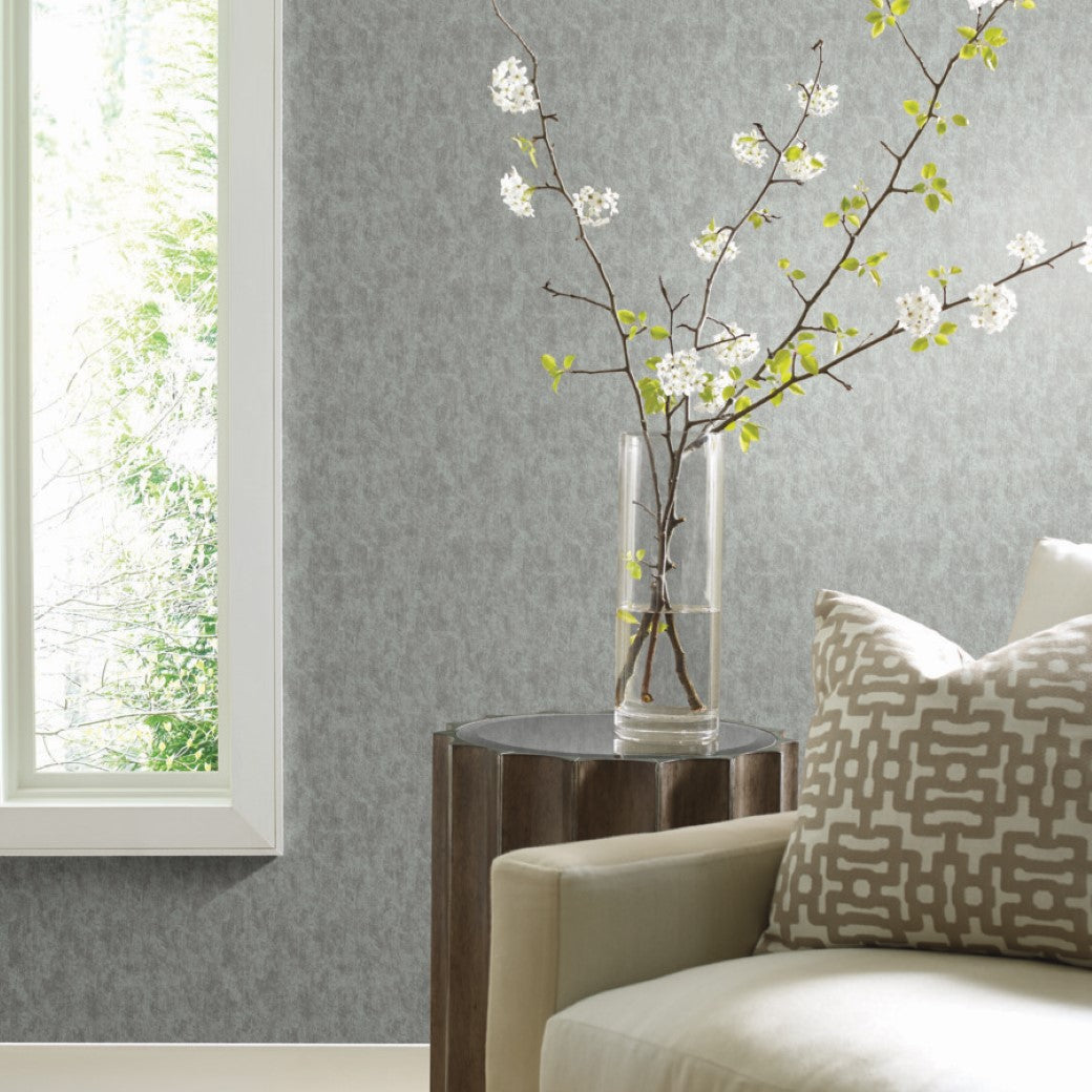 In a contemporary living room corner, a pale armchair with a patterned pillow sits beside a small wooden table holding a glass vase with flowering branches. The background features York Wallcoverings' Spa & Silver Shimmering Patina Wallpaper, enhancing the view of lush greenery outside the window.