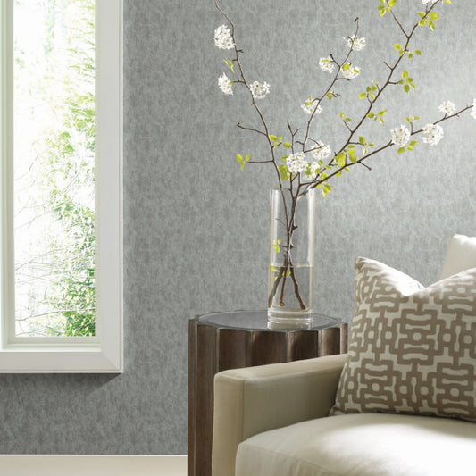 In a contemporary living room corner, a pale armchair with a patterned pillow sits beside a small wooden table holding a glass vase with flowering branches. The background features York Wallcoverings' Spa & Silver Shimmering Patina Wallpaper, enhancing the view of lush greenery outside the window.