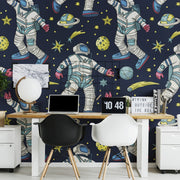 Home Office - A vibrant home office with a wall covered in Decor2Go Wallpaper Mural featuring astronauts and planets. The desk holds a laptop, a small globe, and a decorative sign, flanked by two modern chairs.