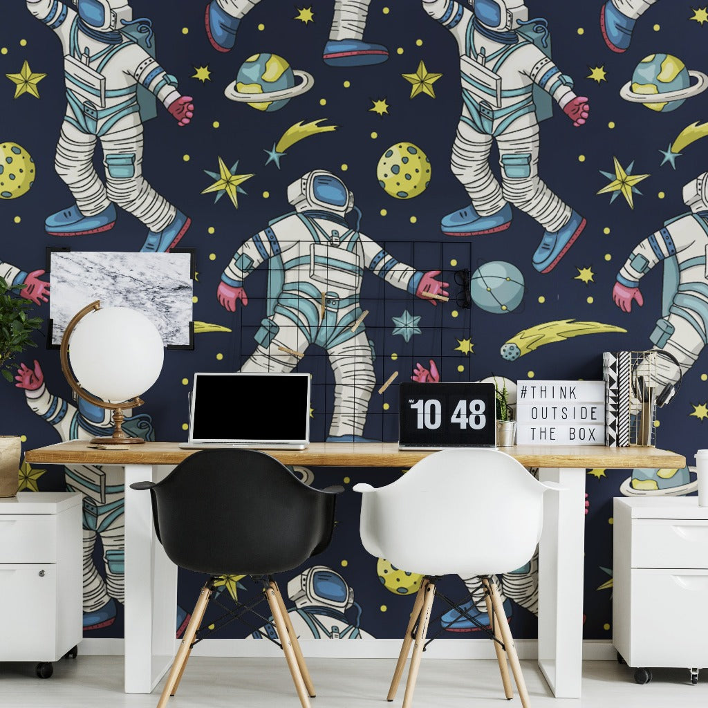 A vibrant home office with a wall covered in Decor2Go Wallpaper Mural featuring astronauts and planets. The desk holds a laptop, a small globe, and a decorative sign, flanked by two modern chairs.
