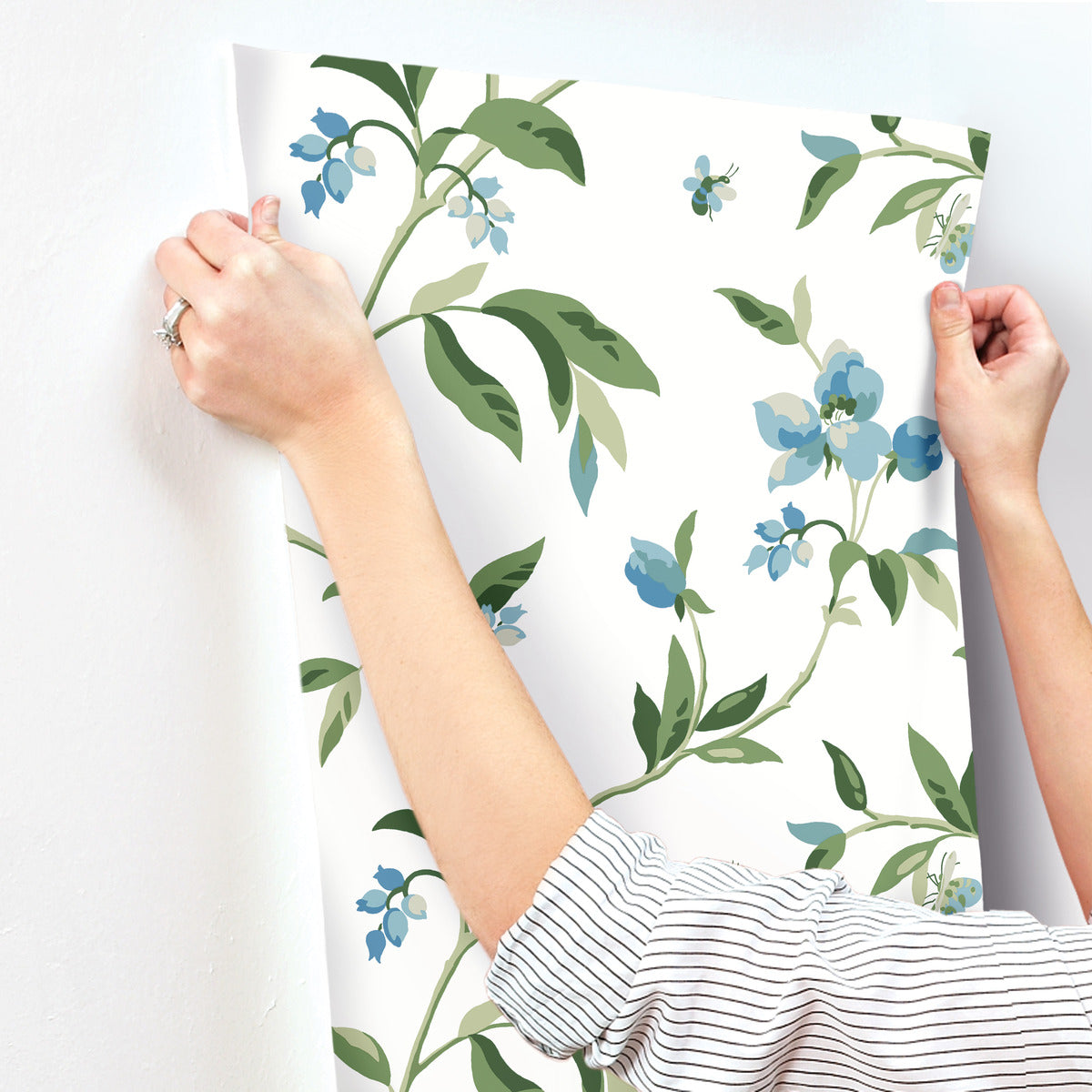 A person hangs a sheet of whimsical floral-patterned wallpaper, featuring green leaves and blue flowers, on a white wall. They wear a striped shirt and a ring on their left hand, showcasing the easy installation of the York Wallcoverings Springtime Midnight/Multi Wallpaper Black, Blue (60 Sq.Ft.).