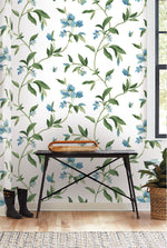 A room with white walls featuring whimsical Springtime Midnight/Multi Wallpaper Black, Blue (60 Sq.Ft.) by York Wallcoverings showcasing green leafy and blue floral patterns. There is a black metal table with a wooden top holding a tray, a plant on the floor to the right, a pair of black boots to the left, and a window letting in natural light.