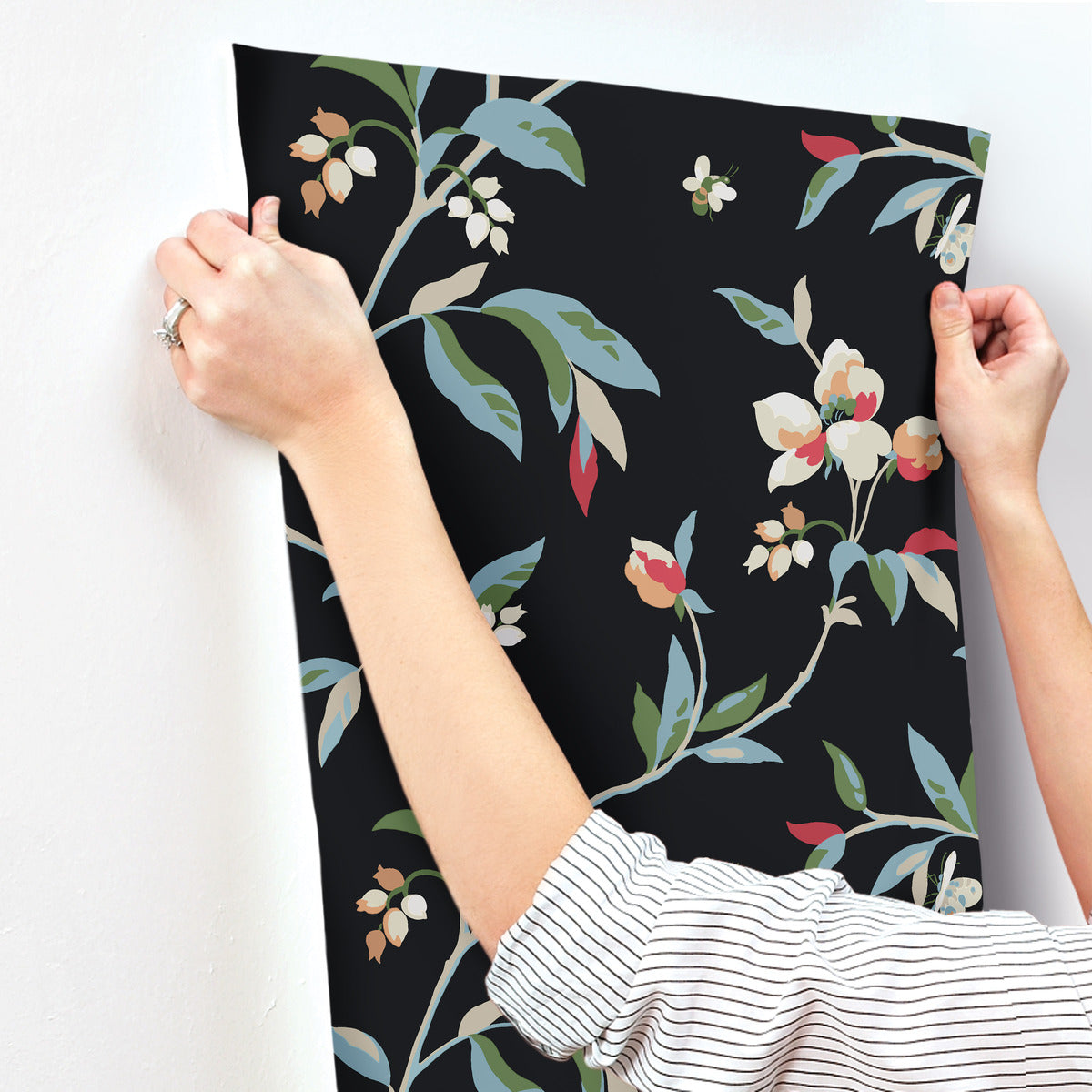 Person in a striped shirt hangs York Wallcoverings' Springtime Midnight/Multi Wallpaper Black, Blue (60 Sq.Ft.) on a white wall. The wallpaper features a black background with colorful flowers and leaves, ensuring easy installation while bringing vibrant floral charm to the room.