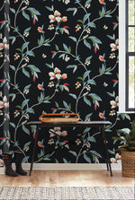 The image showcases a York Wallcoverings Springtime Midnight/Multi Wallpaper Black, Blue (60 Sq.Ft.) with a whimsical design of white flowers, green leaves, and small red buds. A wooden table with black legs and a woven basket on top stands against the wallpaper. Off to the side, you’ll find a pair of black rain boots and a lush green plant.