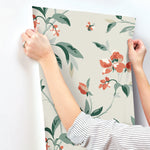 A person wearing a striped shirt is applying York Wallcoverings Springtime Midnight/Multi Wallpaper Black, Blue (60 Sq.Ft.) to a white wall. The whimsical design features large flowers, leaves, and buds. The person's hands are visible, holding the edges of the wallpaper in place for easy installation.