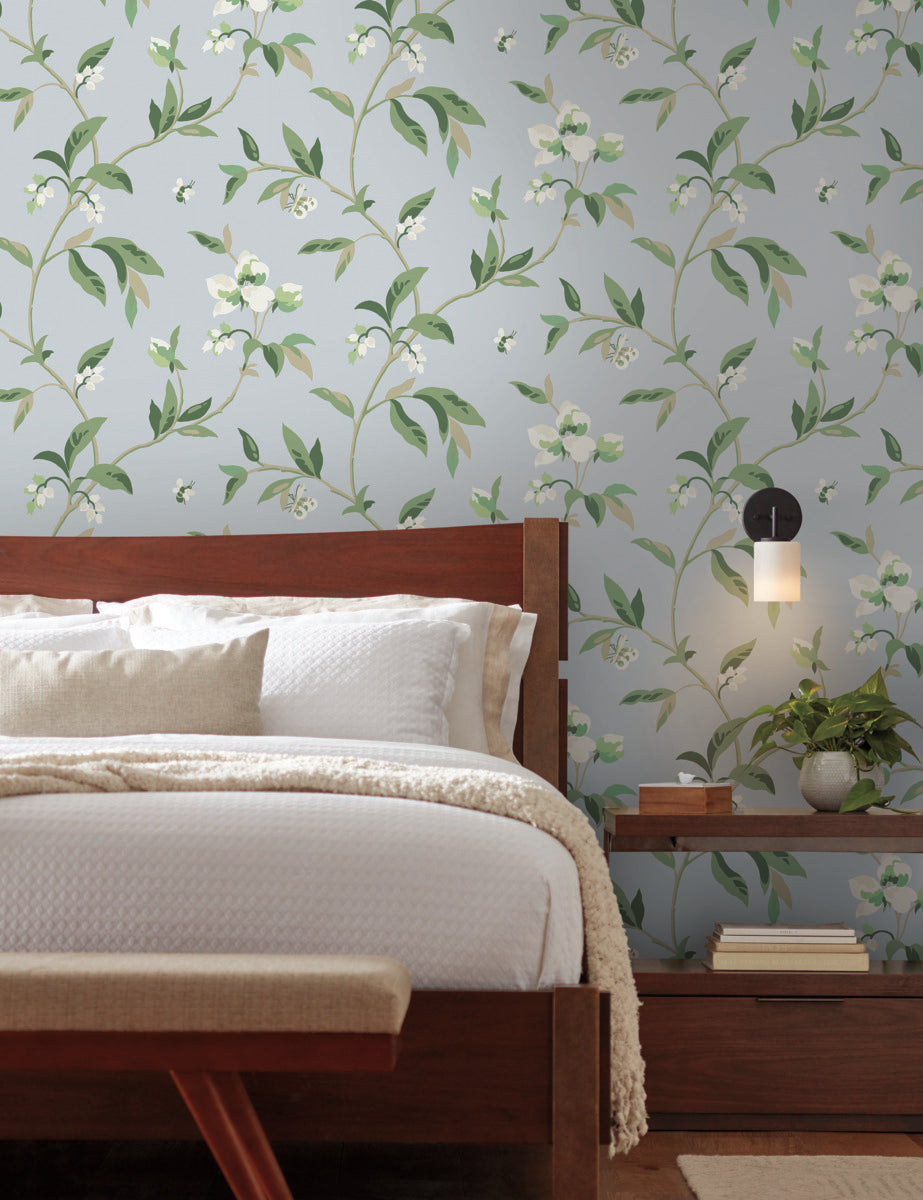 A cozy bedroom with a wooden bed frame and white bedding. A small, round, wall-mounted light fixture is on the right side above a wooden nightstand that holds a few books. The walls boast **York Wallcoverings' Springtime Midnight/Multi Wallpaper Black, Blue (60 Sq.Ft.)** featuring a whimsical design with green leaves and white flowers, ensuring easy installation.