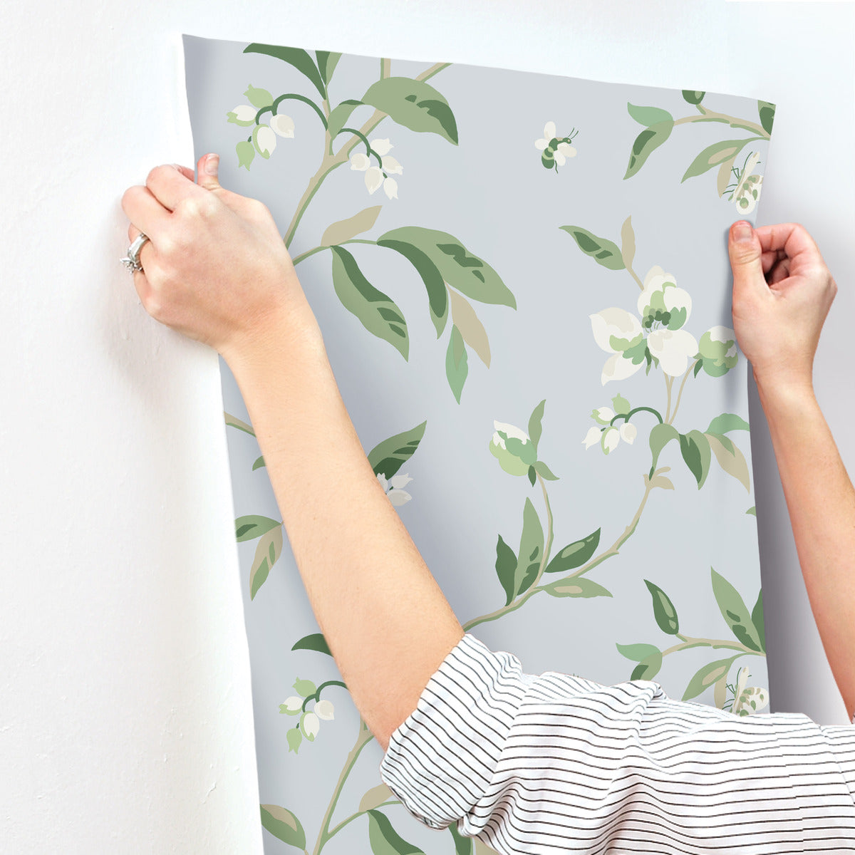 A person with a striped shirt is using both hands to apply a sheet of York Wallcoverings' Springtime Midnight/Multi Wallpaper Black, Blue (60 Sq.Ft.) adorned with a soft white and green botanical pattern to a white wall. The whimsical design showcases delicate flowers and leaves, promising an easy installation for any decor enthusiast.