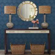 Entryway - A stylish is highlighted by a dark wood console table with a white top, featuring two wicker baskets below and two elegant lamps above. A round mirror with a gold frame enhances the space, reflecting the Starlight Wallpaper in Blue/Gold by York Wallcoverings, adorned with metallic dots. A vase filled with pink flowers rests gracefully on the table
