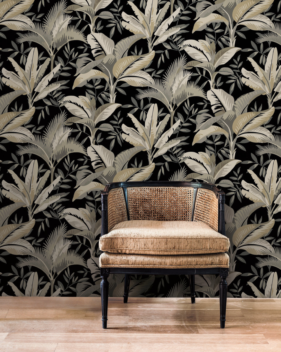 A rattan chair with a black wooden frame sits on a light hardwood floor. The background features the Summerhouse Peacock Wallpaper Blue, Green (60 Sq.Ft.) by York Wallcoverings, creating a stylish and modern interior design that's perfect for tropical living.