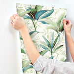 A person with a striped shirt is holding up a sheet of Summerhouse Midnight Wallpaper Black, Brown (60 Sq.Ft.) by York Wallcoverings against a white wall. The easy removable wallpaper features green, blue, and teal leaves on a light background, perfect for adding a touch of waterfront luxury to any room.