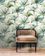 A woven rattan armchair with a dark wooden frame sits on a light wooden floor against a wall covered in York Wallcoverings' Summerhouse Peacock Wallpaper Blue, Green (60 Sq.Ft.), featuring tropical leaf patterns in shades of green and white, creating the perfect tropical living ambiance.