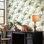 A stylish room featuring a large black desk against York Wallcoverings' Summerhouse Midnight Wallpaper in Orange and Green, with leaf patterns, complemented by a vintage lamp, books, and a small plant on the desk, near an open window.