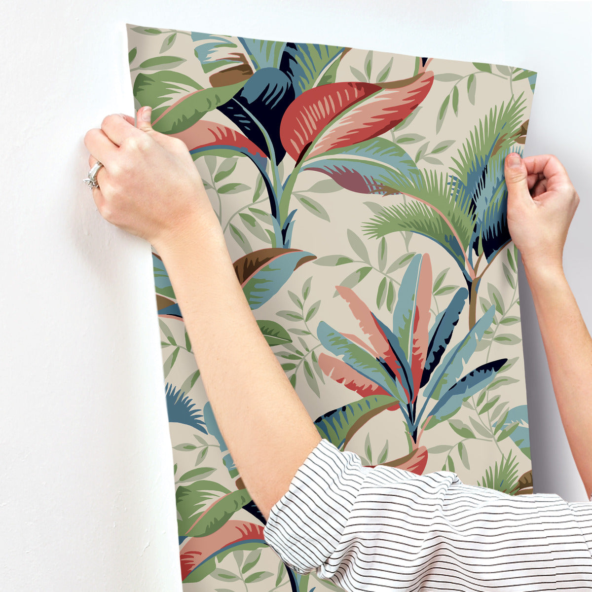 Two hands pinning a York Wallcoverings Summerhouse Midnight Wallpaper Orange, Green (60 Sq.Ft.) on a white wall. The person wears a striped shirt and is adjusting the wallpaper's position.