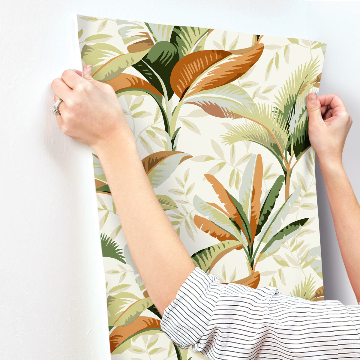 A person wearing a striped shirt is hanging up York Wallcoverings' Summerhouse Midnight Wallpaper Black, Brown (60 Sq.Ft.) featuring vibrant green, brown, and orange leaves. The individual's hands and part of their arms are visible, pressing the easy removable wallpaper onto a white wall.