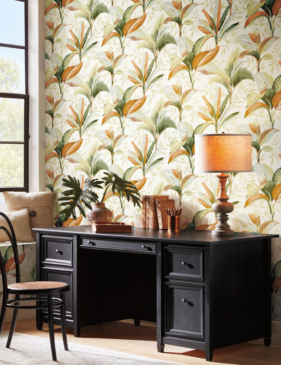 A cozy room corner featuring a black wooden desk with a lamp and plant decorations, set against vibrant Summerhouse Midnight Wallpaper Orange, Green (60 Sq.Ft.) by York Wallcoverings with tropical leaf patterns. A window and a bentwood chair are also visible.