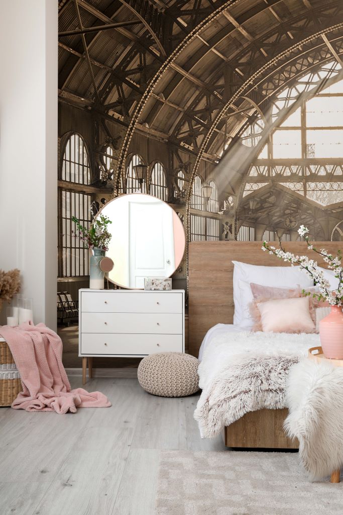 A cozy bedroom set in a spacious room with an industrial-style ceiling and Decor2Go Wallpaper Mural. Furnishings include a fluffy bed, a white dresser with a round mirror, knitted pouf, and.