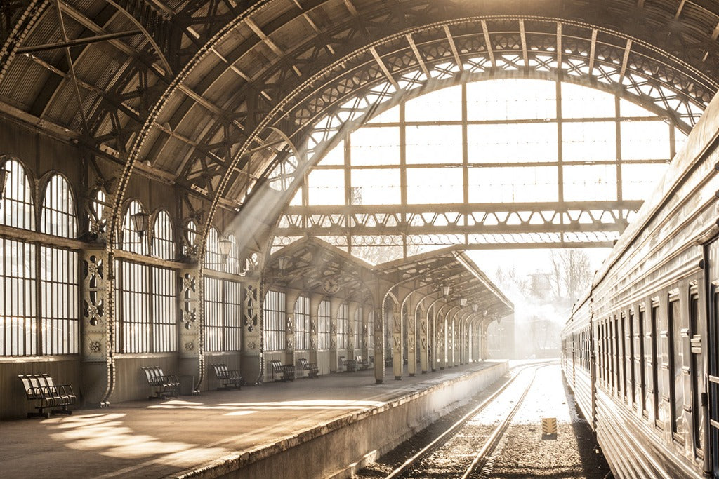Sun Station Wallpaper Mural by Decor2Go streams through the large windows of an ornate, vintage train station, illuminating the platform and an idle train on the side. Dust particles are visible in the air, adding a mystical quality.
