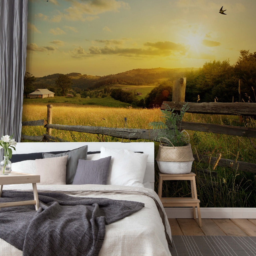 A cozy bedroom with a large Decor2Go Wallpaper Mural of a sunset over a scenic grassy hill with a small farmhouse, featuring a bed with gray and white bedding, and a small table with a vase of