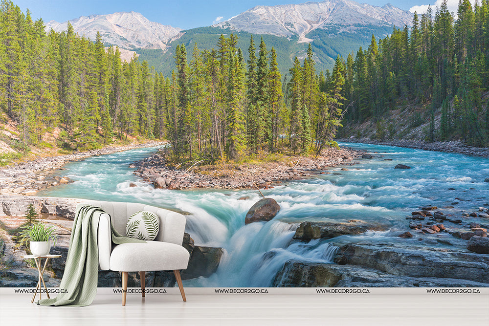 A surreal image of a cozy armchair and a side table with a plant by Sunwapta Falls Wallpaper Mural, flowing through a pine forest with mountains in the background.