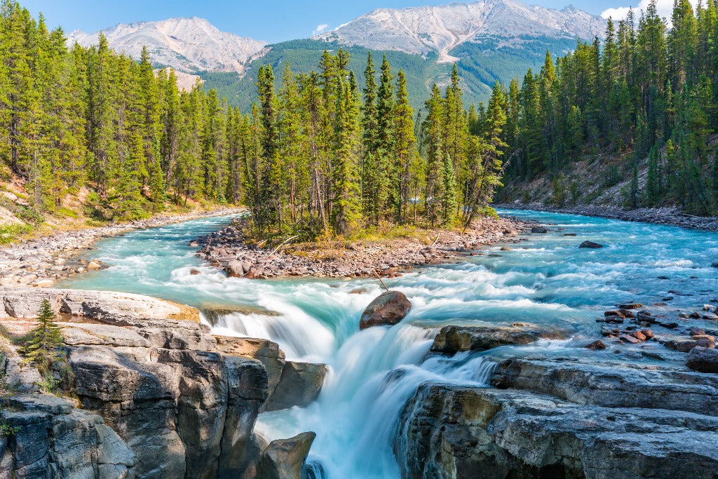 A vibrant river cascades over rocks in a forested mountainous landscape under a clear blue sky, creating the perfect scene for Decor2Go Wallpaper Mural.