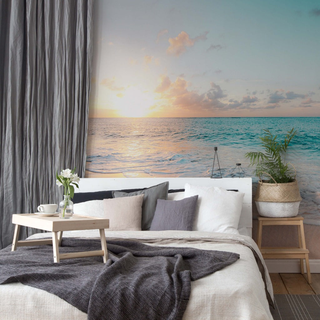 A serene beach-themed bedroom with a large Decor2Go Wallpaper Mural of a sunset over a turquoise sea. A cozy bed is complemented by a small wooden table with a vase of flowers.