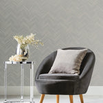 A modern living room features a gray upholstered chair with a cushion, beside which stands a small round glass table from the Modern Metals Collection, displaying a decorative vase with flowers. The Taupe & Silver Polished Chevron Wallpaper by York Wallcoverings adds geometric elegance to the wall.