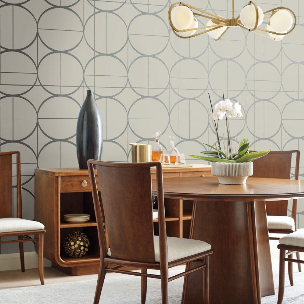 A dining room features a round wood table with cushioned chairs, a Modern Metals Collection sideboard for decor, and a metallic-finish modern chandelier. York Wallcoverings' Taupe & Silver Sun Circles Wallpaper adorns the walls, beautifully complementing the orchid centerpiece with its large circle pattern.