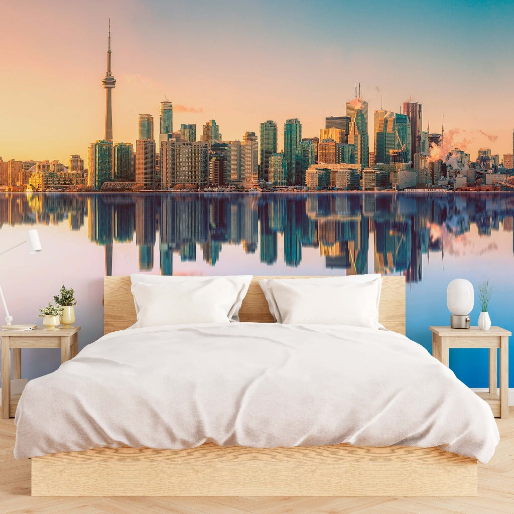 A modern bedroom with a large bed facing a floor-to-ceiling Decor2Go Wallpaper Mural of the Toronto Skyline reflected over a calm body of water at sunset.
