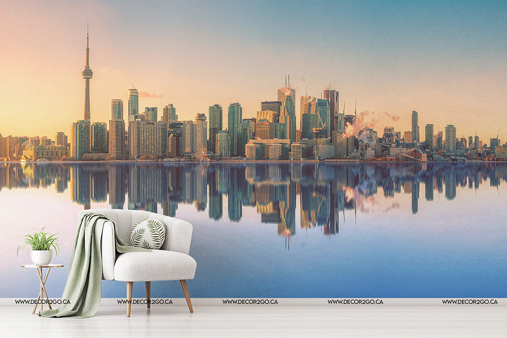 A stylish interior setup with a white armchair and a green throw blanket beside a small plant, with a panoramic backdrop of Toronto's skyline mirrored in calm waters at sunrise, enhanced by a Decor2Go Wallpaper Mural.
