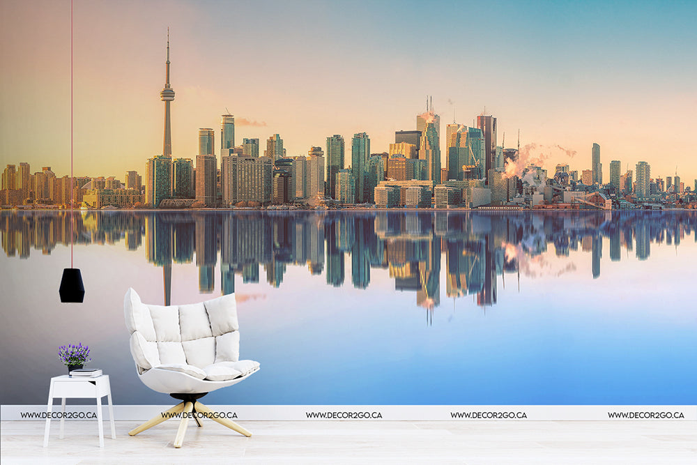 A serene scene of a Decor2Go Toronto Skyline Wallpaper Mural reflected in water, with the CN Tower visible, juxtaposed with an interior setting featuring a stylish white chair and a small table with flowers.