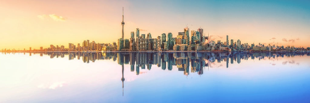 Panoramic view of a city skyline at sunset with the reflections of skyscrapers vividly mirrored in the calm water below, showcasing a vibrant mix of blue and orange hues in the sky. This Toronto Skyline Wallpaper Mural by Decor2Go Wallpaper Mural is truly breathtaking.