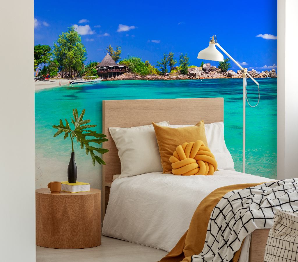 Bedroom - A stylish bedroom with a Decor2Go Tropical Blue Wallpaper Mural on the wall, featuring a wooden bed with beige and yellow pillows, a side table with a lamp and books, and a small plant.